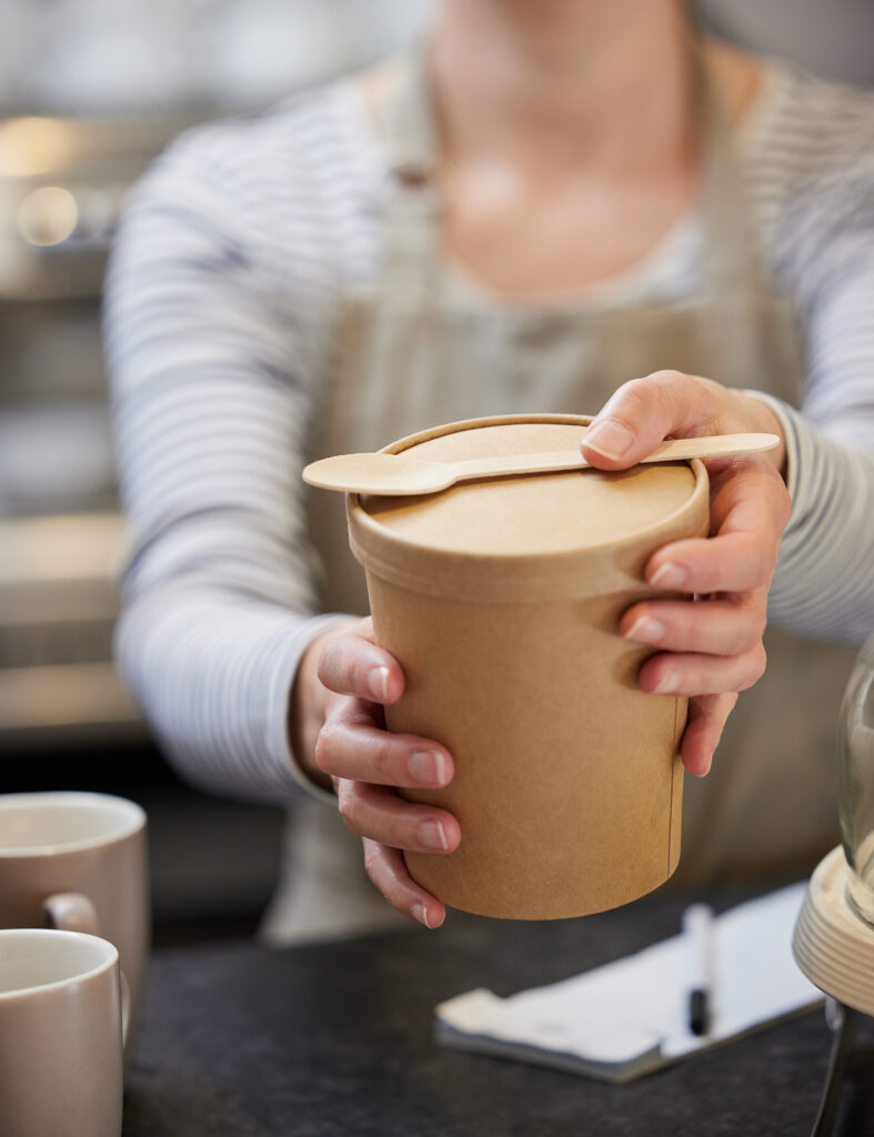 Close Up Of Female Worker in Cafe Serving Meal In Sustainable Recyclable Packaging With Wooden Spoon